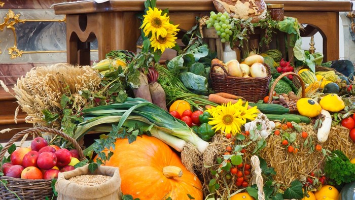 Ein großer Korb gefüllt mit einer Vielzahl von frischem Obst und Gemüse, darunter Kürbisse, Äpfel, Tomaten und Blumen, steht auf einem Tisch.