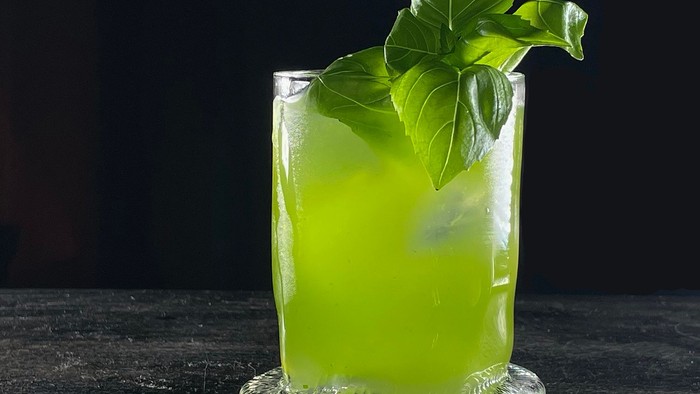 En forfriskende grøn cocktail med basilikumblad som pynt, serveret i et stilfuldt glas på en underkop.