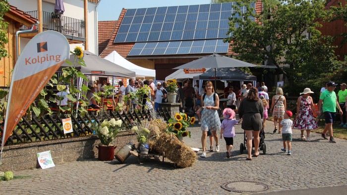 Freiluftmarkt mit Pflanzen, Heuballen und einem solarbetriebenen Gebäude, an dem Familien und Besucher stöbern.