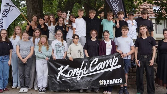 Eine Gruppe Jugendlicher unter einem Baum mit einem Banner mit der aufschrift KonfiCamp
