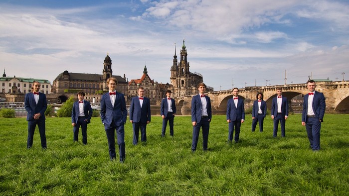 Eine Gruppe von Männern in formellen Anzügen posiert auf üppigem grünem Rasen mit historischer Architektur im Hintergrund.