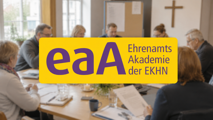 Eine Gruppe von Menschen sitzt gemeinsam an einem Tisch mit Unterlagen; im Hintergrund hängt ein Kreuz an der Wand. Im Vordergrund steht groß das gelb-violette Logo „eaA – EhrenamtsAkademie der EKHN“.