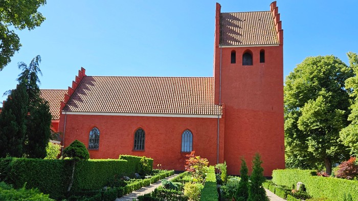 Livligt rødt kirkebyggeri med frodige haver og en klar blå himmel som baggrund.