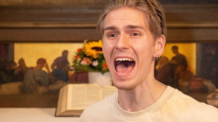 Ein junger Mann jubelt fröhlich vor einer festlichen Innenszene mit einem dekorierten Tisch und einem gemalten Hintergrund.
