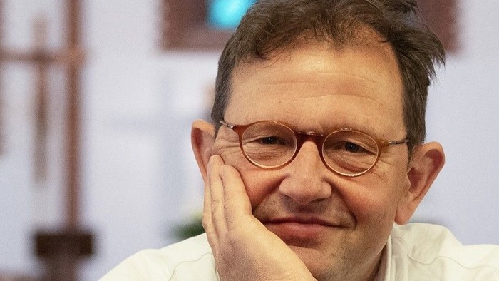 Ein nachdenklicher Mann mit Brille stützt sein Kinn auf die Hand und trägt ein helles Hemd.