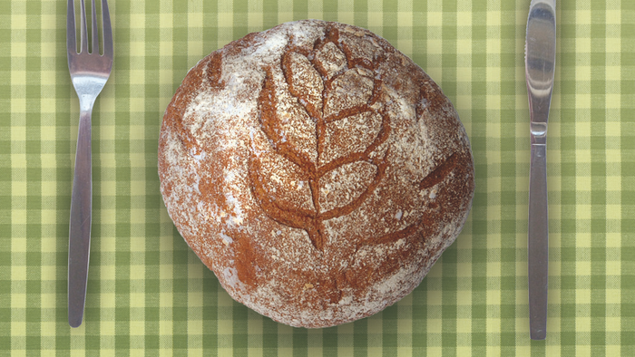 Handgefertigtes Brot mit Blattprägung oben, mit Mehl bestäubt, auf einem karierten Tischtuch mit Besteck platziert.