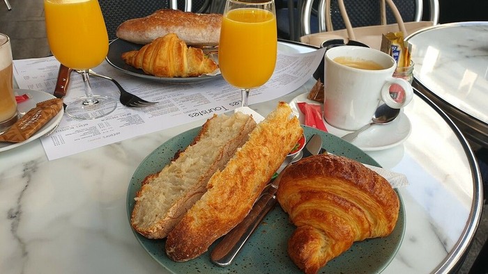Frühstückstisch im Restaurant: Zwei Teller mit jeweils einem angerösteten Brötchen und einem Croissant und einem vollen Glas Orangensaft stehen auf einem Tisch. Dazu Kaffeee.