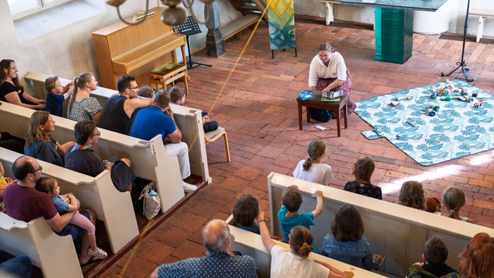 Kinder und Erwachsene sitzen während der Kirche Kunterbunt in den Kirchenbänken. Vor dem Altar erzählt eine Mitarbeiterin eine Geschichte, vor ihr liegen Figuren und Materialien auf einer Decke.