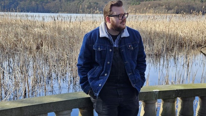Jugendmitarbeiter Friedrich Wolter auf einer Brücke, im Hintergrund ein See mit Schilfpflanzen. Dunkle Kleidung, darüber blaue Jeansjacke mit weißem Kragen. Bart, dicke schwarze Brille, kurze, braune Haare.