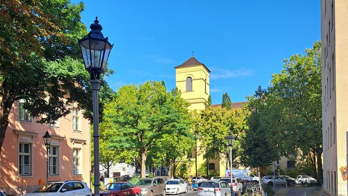 Eine malerische Straßenszene mit geparkten Autos, üppigen Bäumen und einem historischen Kirchturm unter klarem blauem Himmel.