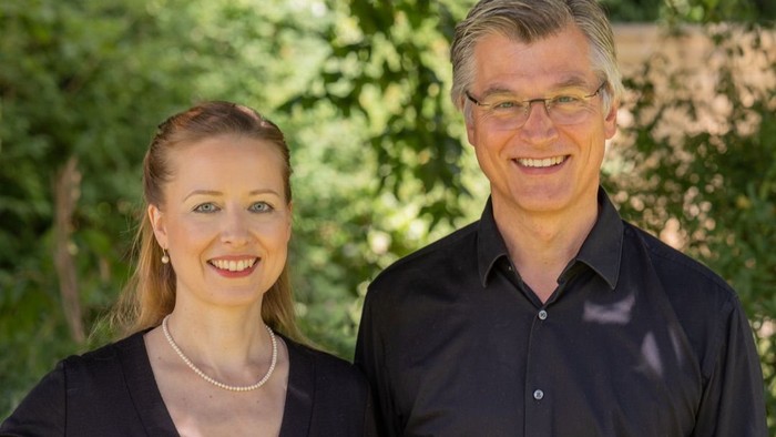 Ein Mann und eine Frau lächeln in die Kamera, vor grünem Blattwerk.