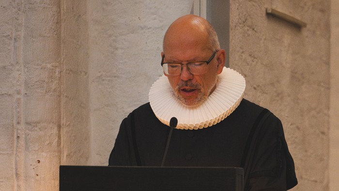 Ein glatzköpfiger Mann in akademischer Robe spricht an einem Rednerpult in einem formellen Innenraum.