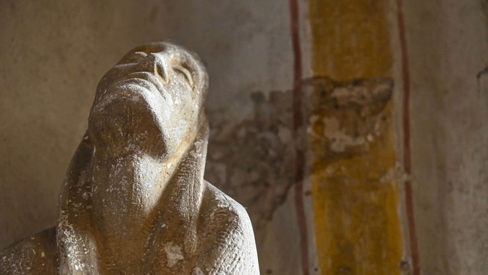Verwitterte Statue mit vor dem Gesicht gefalteten Händen in einem verfallenen, antiken Innenraum.