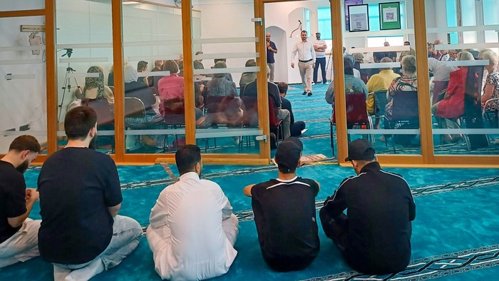 Ein Blick in einen modernen Moscheeraum. Im Vordergrund fünf junge Männer auf dem Boden. Vor ihnen eine Glasswand. Dahinter eine gemischte Gruppe auf Stühlen.