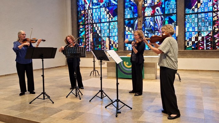 Vier Musiker spielen Saiteninstrumente und singen in einem Raum mit bunten Kirchenfenstern.