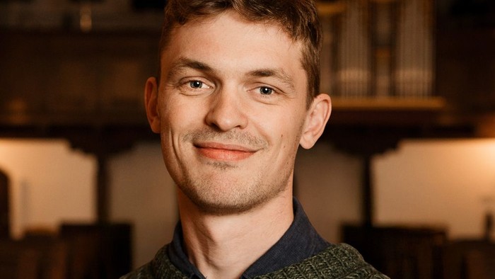 En smilende mand i en grøn sweater står foran en rustik træbøjle i baggrunden.