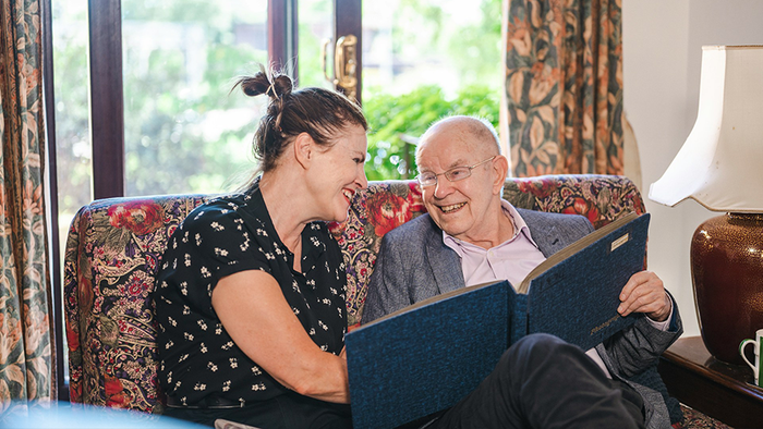 Lächelnder alterer Mann sitzt mit Frau auf Couch und zeigt ihr ein Fotoalbum.