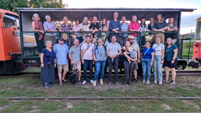Eine Gruppe von rund 25 Erwachsenen und einem Kind steht und sitzt vor einem offenen Wagen einer Schmalspurbahn; links und rechts sind Teile zweier historischer Triebfahrzeuge zu sehen.
