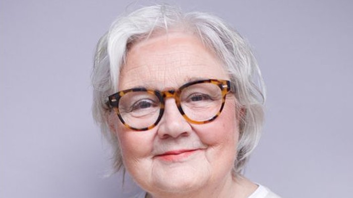 Elegant ældre kvinde med skildpaddebriller og en hvid top, der smiler blidt.