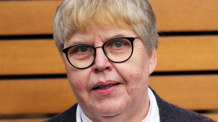 Eine ältere Frau mit kurzem grauen Haar und Brille trägt ein graues Pullover mit weißem Kragen.