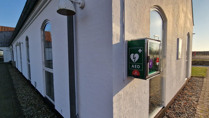 Et udendørsbillede af en bygning med en automatisk ekstern defibrillator (AED) monteret på væggen ved siden af en buet døråbning.