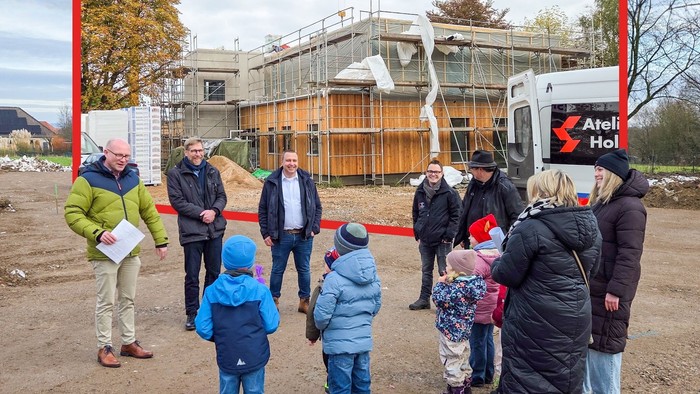Ein Kompositbild: Im Vordergrund eine Gruppe Kinder und Erwachsene auf einer leeren Baustelle. Dahinter im Rahmen ein fast fertiger Neubau mit Holzverkleidung