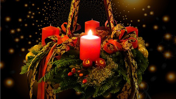 Ein Adventskranz mit vier Kerzen, geschmückt mit roten Bändern und Tannenzapfen, vor einem dunklen Hintergrund mit goldenen Lichtern.