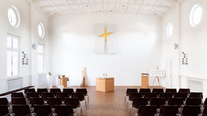 Heller Kirchenraum mit Reihen dunkler Stühle, einem schlichten Altar aus Holz und einem großen weißen Kreuz mit goldfarbenem Hintergrund an der Stirnwand. Rundfenster lassen viel Tageslicht in den Raum.