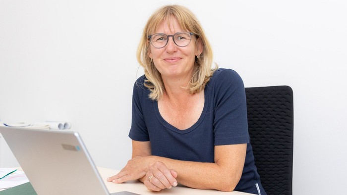 Eine lächelnde Frau mit Brille arbeitet an einem Laptop an einem Schreibtisch, neben dem ein Notizbuch liegt.