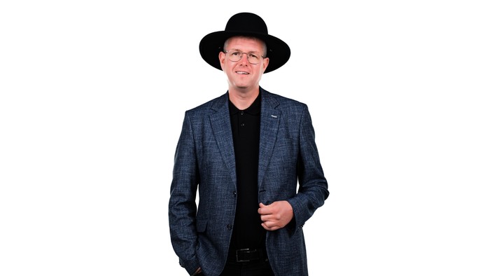 En mand i en grå tweedjakke, sort skjorte og fedora poser selvsikkert med hænderne i lommerne.