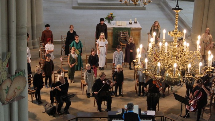 Ein Chor und Musiker treten in einer prächtigen Kirche mit kunstvollen Kerzenkronleuchtern und historischer Architektur auf.