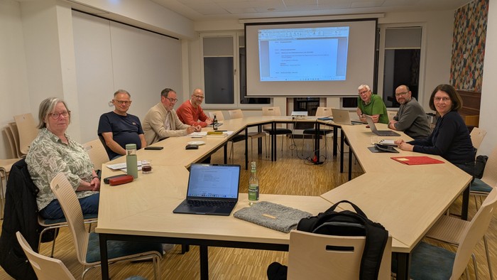 Eine Gruppe von Personen versammelte sich um einen Konferenztisch in einem Besprechungsraum mit einer Präsentation auf dem Bildschirm.