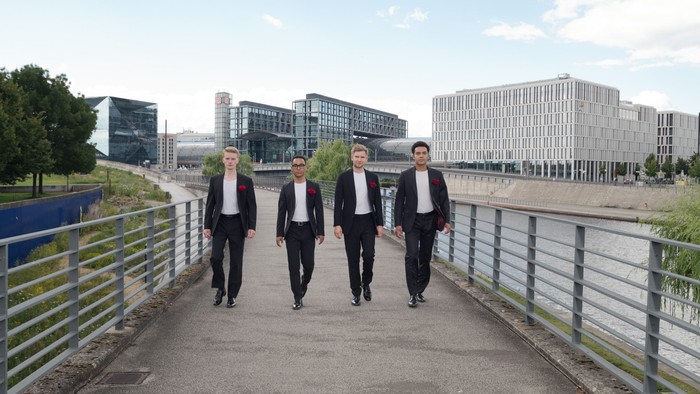 Vier Männer in formeller Kleidung gehen gemeinsam über eine moderne Brücke in der Nähe zeitgenössischer Gebäude.