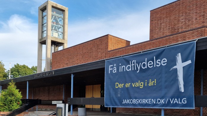 Kirkebygningens indgang med parkerede cykler og et skilt, der opfordrer til fællesskabets deltagelse i valget.