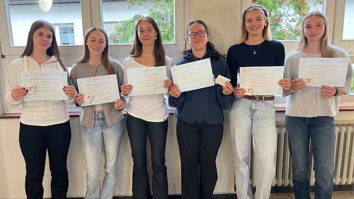 Fünf Studierende halten stolz ihre Zertifikate in einem hellen Innenflur mit großen Fenstern.