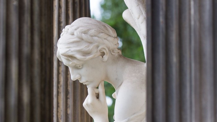 Eine klassische Marmorstatue einer nachdenklichen Figur, die ihr Kinn auf die Hand stützt, zwischen Säulen.