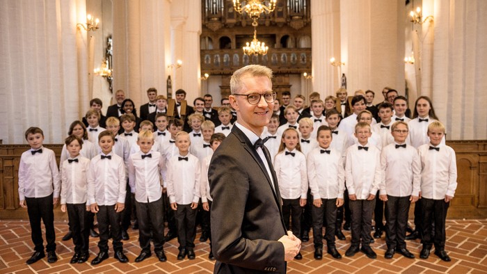 En dirigent poser med en gruppe unge mandlige kormedlemmer i en prægtig kirkesal.