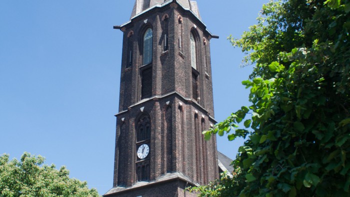 Ein hoher Kirchturm aus Ziegeln mit Uhr und spitzem Dach, umgeben von Bäumen und Büschen.