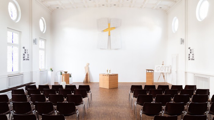 Heller Kirchenraum mit Reihen dunkler Stühle, einem schlichten Altar aus Holz und einem großen weißen Kreuz mit goldfarbenem Hintergrund an der Stirnwand. Rundfenster lassen viel Tageslicht in den Raum.