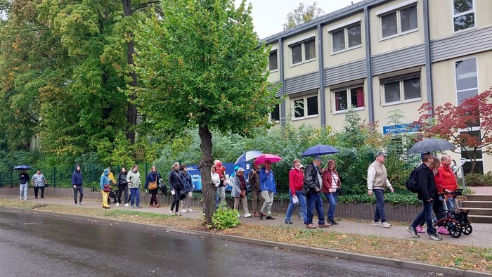 Menschen warten unter Regenschirmen auf einem nassen Gehweg in der Nähe eines Gebäudes und von Bäumen.
