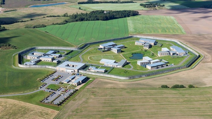 Sikret anlæg omgivet af hegn i et landligt, grønt landskab.