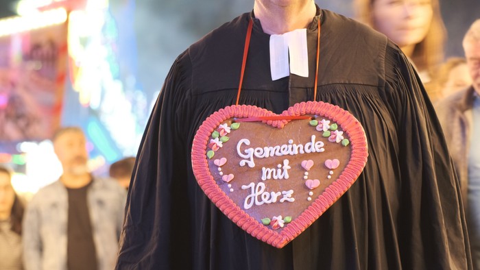 Eine Person in geistlicher Kleidung hält ein herzförmiges Lebkuchen-Schild mit der Aufschrift *„Gemeinde mit Herz“*.