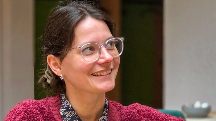 Eine junge Frau mit dunklen Haaren und Brille sitzt mit einer Tasse an einem Tisch