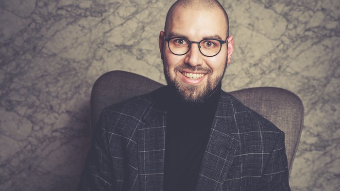 Ein bärtiger Mann mit Brille und einem karierten Sakko lächelt selbstbewusst in einem gemütlichen Sessel.