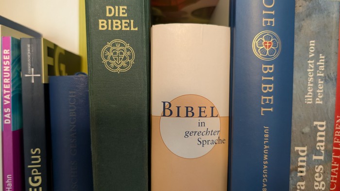 Die „Bibel in gerechter Sprache“ steht in einem Bücherregal zwischen einer grünen und einer blauen Bibelausgabe. Auf ihrem hellen Buchrücken ist der Titel gut lesbar.