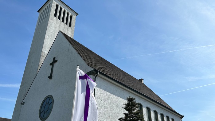 Moderne Kirche mit weißer Fassade, hohem Kirchturm und einer lila-weißen Trauerflagge, die außen angebracht ist.