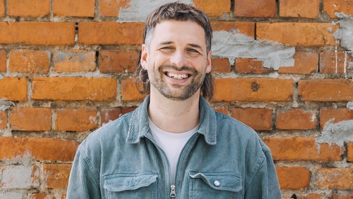 Ein fröhlicher Mann mit langen Haaren lächelt vor einer rustikalen Backsteinmauer, er trägt ein Denim-Hemd über einem weißen T-Shirt.