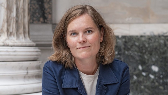 En person i en blå jakke og et mønstret nederdel sidder eftertænksomt på en hvid bænk udendørs.
