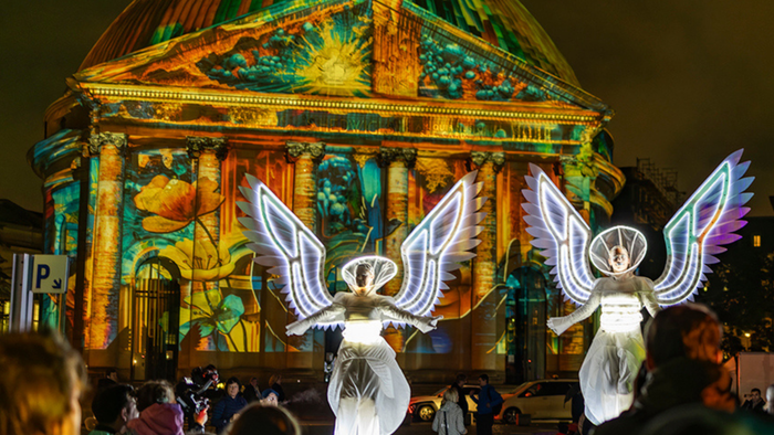 Bunte Lichtprojektionen erleuchten nachts ein prächtiges Gebäude, während in der Nähe engelsgleiche Figuren leuchten.