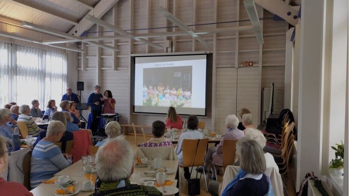 Die Gruppe versammelt sich drinnen für eine Präsentation mit gedeckten Tischen für eine Mahlzeit und einem projizierten Bild auf der Leinwand.